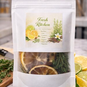 May include: A white bag with a clear window displaying dried citrus slices, herbs, and spices. The bag features a label with the text "Fresh Kitchen" and ingredients like lime, cinnamon, and oregano. Fresh lemons and limes are in the background.
