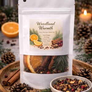 May include: A white pouch labeled "Woodland Warmth" with ingredients for a simmer pot. The pouch contains dried oranges, cinnamon sticks, pine sprigs, cloves, and cranberries. The ingredients are displayed on a wooden tray with pine cones and a bowl of simmer pot ingredients.