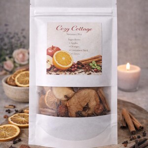 May include: A white pouch labeled "Cozy Cottage" with a clear window revealing dried fruit and spices. The label lists ingredients: apples, oranges, cinnamon sticks, and cloves. A candle and sliced oranges are in the background.
