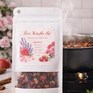 May include: A white bag labeled "Love Is in the Air" with a clear window showing a Valentine's Day simmer pot mix. The label lists ingredients like strawberry, cherries, and rose petals. Steam rises from a pot in the background.