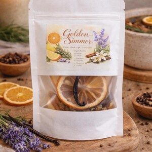 Puede incluir: Una bolsa blanca con una ventana transparente, llena de rodajas de cítricos secos, vainilla y especias. La etiqueta dice "Golden Simmer" con los ingredientes enumerados. La imagen incluye una vela, lavanda y cuencos de madera.