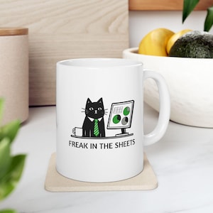 May include: White ceramic mug with a black cat wearing a green tie and sitting at a computer with a chart on the screen. The text "Freak in the sheets" is printed below the cat.