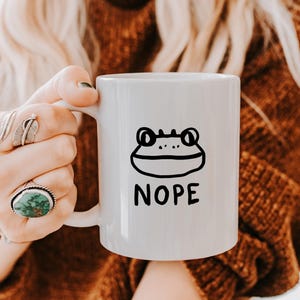 以下が含まれることがあります： 白地に黒と白の蛙のイラストと「NOPE」の文字が印刷された白いセラミックマグカップです。
