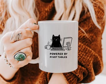 Könnte beinhalten: Weiße Keramiktasse mit einer schwarzen Katze, die an einem Computer sitzt, und dem Text "Powered by Pivot Tables".
