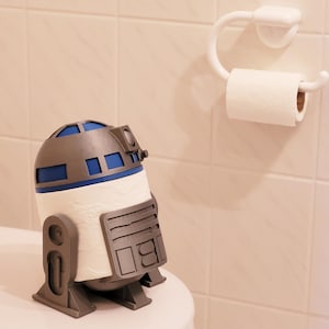 Peut inclure: Un porte-papier toilette en forme de R2-D2 blanc, bleu et gris. Le support est posé sur une surface blanche à côté d'un rouleau et d'un support de papier toilette blancs. La salle de bain est carrelée de blanc.