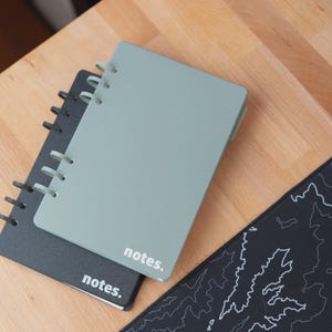 Può includere: Due quaderni a spirale, uno grigio-verde e uno nero, sono impilati su una superficie di legno. Il quaderno grigio-verde ha la parola "notes." stampata sulla copertina. È visibile anche un tappetino per mouse nero con un disegno topografico bianco.