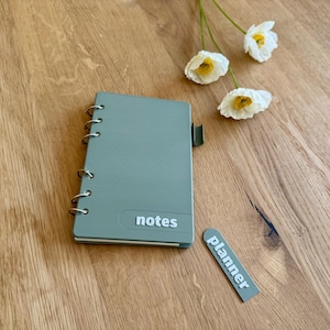May include: A sage green notebook with silver ring binding and the word "notes" printed on the cover. A small "planner" label is next to the notebook. White and yellow flowers are on a wooden surface.