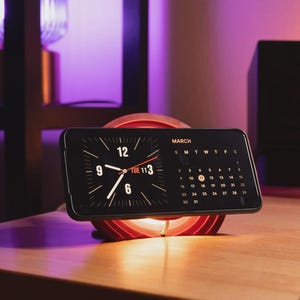 Könnte beinhalten: Ein schwarzes Smartphone mit Uhrzeit und Kalender, das auf einem roten, kreisförmigen Ständer liegt. Die Uhr zeigt 9:07 Uhr, Dienstag, 11. März. Der Ständer steht auf einem Holzschreibtisch, im Hintergrund ein Lautsprecher und andere Gegenstände.