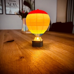 Puede incluir: Una pequeña luz nocturna de globo aerostático iluminada con un patrón de rayas rojas, naranjas, amarillas y azules. El globo está sobre una mesa de madera.