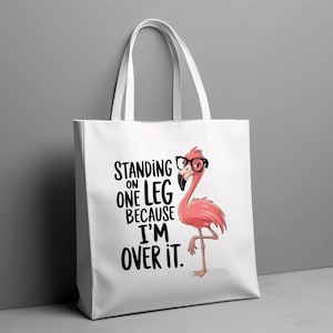 May include: White tote bag featuring a pink flamingo wearing glasses and the text "Standing on one leg because I'm over it."