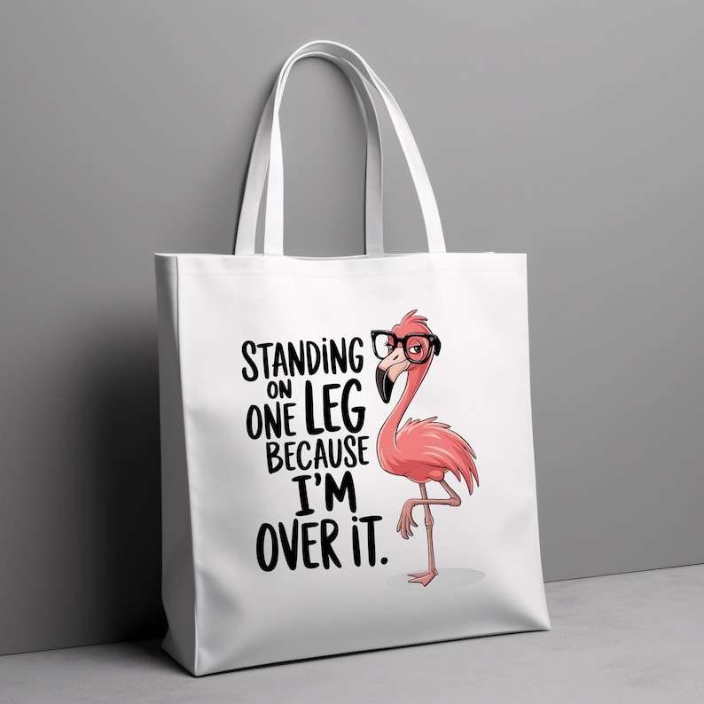May include: White tote bag featuring a pink flamingo wearing glasses and the text "Standing on one leg because I'm over it."
