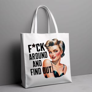 Puede incluir: Bolso tote blanco con una ilustraci&oacute;n de estilo vintage de una mujer con cabello rubio y el texto "F*ck Around And Find Out" en negro. La mujer lleva un top negro. El bolso tiene asas blancas.