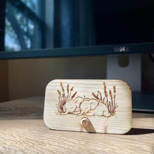 May include: A wooden sign with a laser-engraved design of two elephants sleeping in a field of tall grass. The sign has a small wooden knob on the bottom.