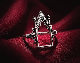 May include: A silver-toned metal ring featuring a structural design that mimics a pointed gothic archway. The ring has a thin band and an open rectangular center framed by intricate architectural details including small spires at the top corners.