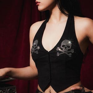 May include: A black halter-neck waistcoat featuring a deep V-neckline and a front button fastening. The garment is embellished with two prominent skull and crossbones motifs created from small, reflective silver-toned rhinestones on each chest panel.