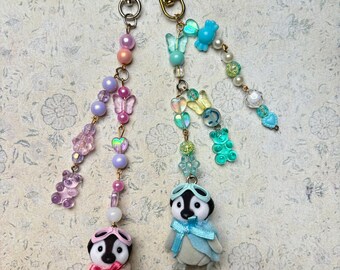 May include: Two penguin keychains with colorful beaded chains. The penguins are wearing aviator goggles and have a pink and blue scarf. The chains have butterflies, hearts, and other charms.