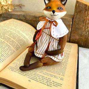 May include: A handmade fox doll wearing a brown and white striped dress with a red heart on the front. The doll is sitting on an open book with a brown cover.