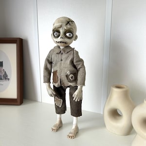 May include: A doll with a pale, textured head featuring large white eyes, dark eye sockets, and stitched mouth details. It wears a tan collared shirt with a dark patch and a skull emblem, paired with dark brown corduroy trousers featuring a small patch.