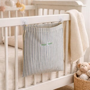 May include: A striped fabric storage bag with the text "BABY BAGS" in green hangs from a white crib. The bag has blue and white vertical stripes. A white blanket and a teddy bear are also visible.