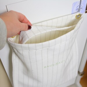 May include: A white fabric storage bag with vertical gray stripes and the text "BAGS RAGS" in green. The bag is hanging on a white cabinet door with a hand reaching into the bag.