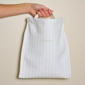 May include: A white canvas bag with vertical gray stripes is held by a hand. The bag has a drawstring closure and the words "BAGS BAGS" are printed in light green. The bag is against a neutral background.