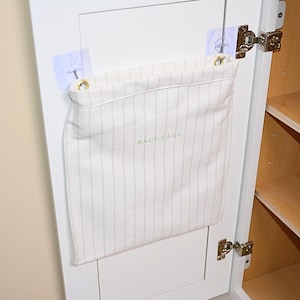 May include: A cream-colored fabric storage bag with vertical pinstripes is attached to a white cabinet door. The bag is secured with clear adhesive hooks and has the words "BAGE BAGS" printed on it. The cabinet interior features wooden shelves.