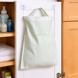 May include: A striped fabric bag with a handle is hanging on a white cabinet door. The bag has vertical stripes in light green and white. The text "BAGS BAGS" is printed on the bag. The cabinet door is attached to a wooden cabinet.