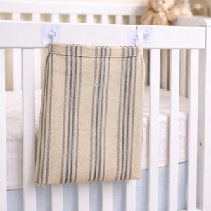 May include: A beige and blue striped fabric storage bag hanging from a white crib. The bag is attached with clear adhesive hooks. The crib has a light blue sheet and a white mattress. A star and a teddy bear are in the background.