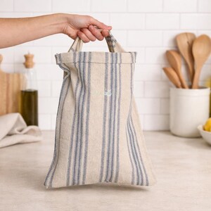 May include: A beige and blue striped tote bag with handles is held up by a person's hand. The bag is made of a textured fabric and has vertical blue stripes. The background includes kitchen items like wooden spoons and a cutting board.