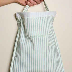 May include: A vertically striped tote bag in white and light blue, held by a hand. The bag has a matching striped handle and the words "BAGS BAGS" embroidered in green.