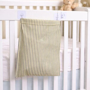 May include: A yellow and gray striped fabric storage bag hanging on a white crib. The bag is attached with clear adhesive hooks. A teddy bear and a star-shaped decoration are visible in the background.