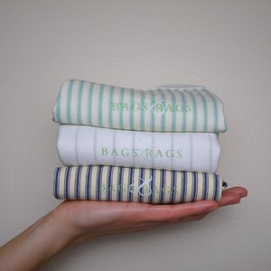 May include: Three rolled fabric items with striped patterns in blue, white, and yellow, held in a hand. The top item is light green with white stripes, the middle is white with thin stripes, and the bottom is blue and yellow. The text "BAGS RAGS" is visible.