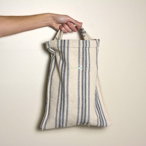 May include: Cream-colored tote bag with vertical navy blue stripes. The bag has a handle and a small embroidered design. The bag is held by a person's hand against a neutral background. The bag is made of a textured fabric.