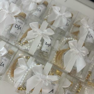 May include: Several clear glass bottles with white labels and gold lids, each containing a clear liquid. The bottles are tied with white ribbon bows and paired with pearl and gold bead necklaces. The labels on the bottles read "EAU DE ROSE" and "DIA".