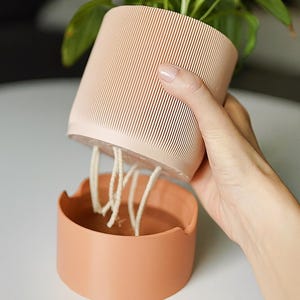 May include: A peach-colored self-watering planter with a ribbed exterior, holding a green plant. The planter is lifted above a matching base, revealing white wicks for water absorption. The design is modern and functional for indoor gardening.