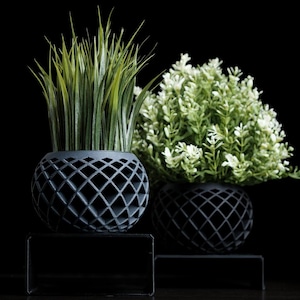 May include: Two decorative planters with faux greenery. The planters are dark gray with a diamond pattern. One holds tall, green grass-like plants, and the other has a bush of small white flowers. Both sit on black stands.