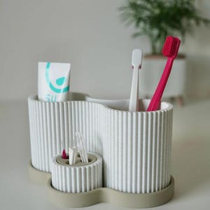 May include: A white, ribbed toothbrush holder with a light gray base, holding a tube of toothpaste, two toothbrushes, and interdental brushes. The holder has a unique, wavy design and is made of a textured material. The toothpaste tube has the word "SU" printed on it.