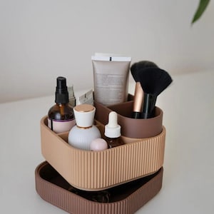 May include: A two-tiered, ribbed, beige and brown cosmetic organizer filled with beauty products. The top tier holds bottles, tubes, and makeup brushes. The bottom tier is a tray. The organizer is designed to hold various cosmetic items.