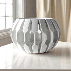 May include: A white, geometric decorative bowl with a spherical shape. The bowl features a series of vertical, angular cutouts, creating a textured surface. The bowl is sitting on a white surface, with a window and curtains in the background.