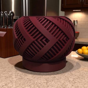 May include: A burgundy-colored decorative bowl with a geometric pattern. The bowl has a rounded shape with a flat base and a textured surface. The design features intersecting lines and angled shapes, creating a woven appearance. The bowl is sitting on a countertop.