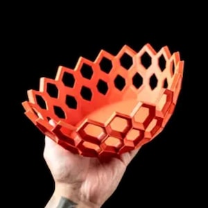 May include: An orange, honeycomb-patterned bowl is held against a black background. The bowl's design features hexagonal cutouts, creating an open, airy aesthetic. The bowl is likely made of plastic or a similar material.