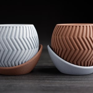 May include: Two decorative ceramic pots with a chevron pattern. One pot is light gray, the other is brown. Each pot sits on a matching saucer. The pots are designed for indoor plants and home decor.