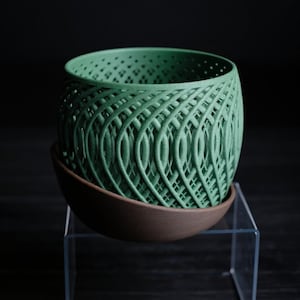 May include: A green, intricately designed planter with a lattice pattern, sitting atop a brown, rounded saucer. The planter has a cylindrical shape with a series of openings. The design is a modern take on a classic plant pot.