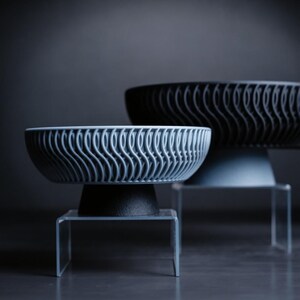 May include: Two decorative bowls with a wave-like pattern, one light blue and one black, sit on clear stands. The bowls have a modern design and are set against a dark gray background.