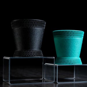 May include: Two decorative black and teal-colored vases with a textured surface and a diamond-patterned mesh design. The vases are displayed on clear acrylic stands against a black background. The vases have a unique, modern design.