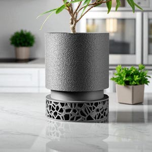 May include: A gray, textured cylindrical planter with a small tree. The planter sits on a decorative base with a geometric pattern. A small green plant is in the background.