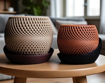 May include: Two decorative planters with intricate woven designs sit on a wooden table. One is beige, the other is terracotta. Each planter rests on a matching saucer, one burgundy, the other black. The planters are a stylish home decor item.