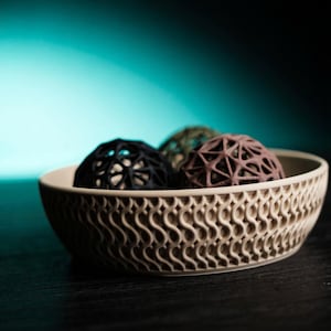 May include: A beige ceramic bowl with a wavy, openwork design holds three decorative spheres. The spheres are black, olive green, and brown, each with a geometric, open pattern. The bowl sits on a dark surface.
