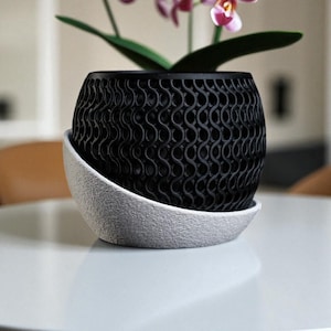 May include: A black, patterned plant pot with a matching white saucer. The pot has a unique, wave-like design. The pot holds a green plant with pink and purple flowers. The pot and saucer are sitting on a white surface.