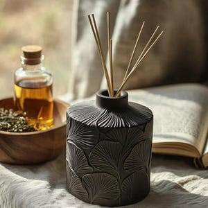May include: A black ceramic essential oil diffuser with a leaf pattern, containing wooden reeds. A small glass bottle with a cork stopper and amber liquid sits nearby, along with a wooden bowl. The items are arranged on a light-colored surface.